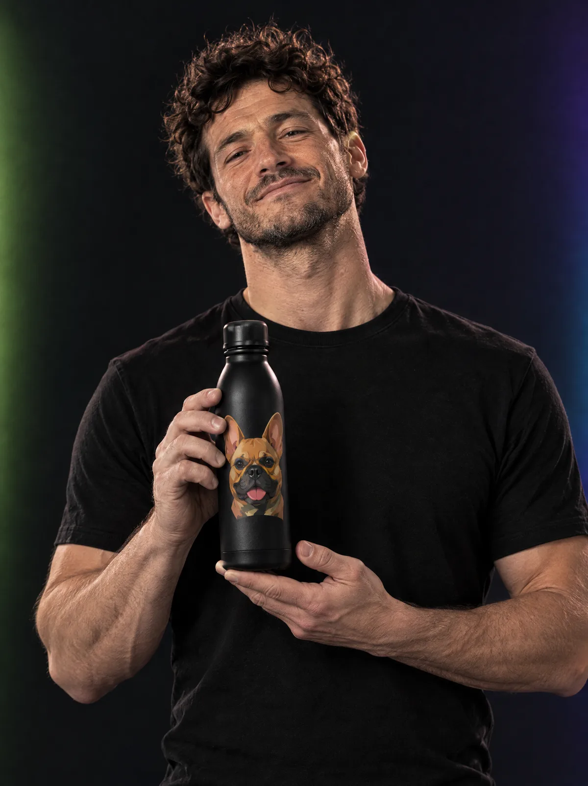 A young man with curly hair in a dark studio lit with coloured gels, holding up a matte black bottle printed with a French bulldog