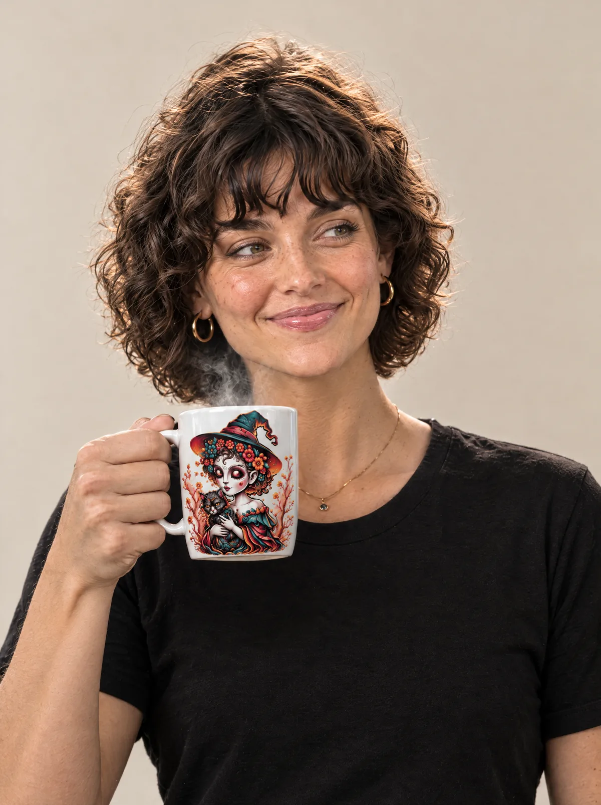 The young woman with curly hair raising a steaming white mug printed with a witch cuddling a black cat